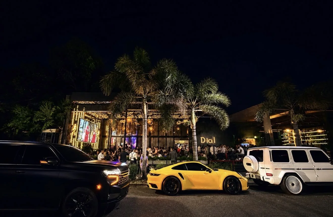 Fachada de Dial Bar & Lounge de noche, con palmeras iluminadas y autos de lujo en la entrada